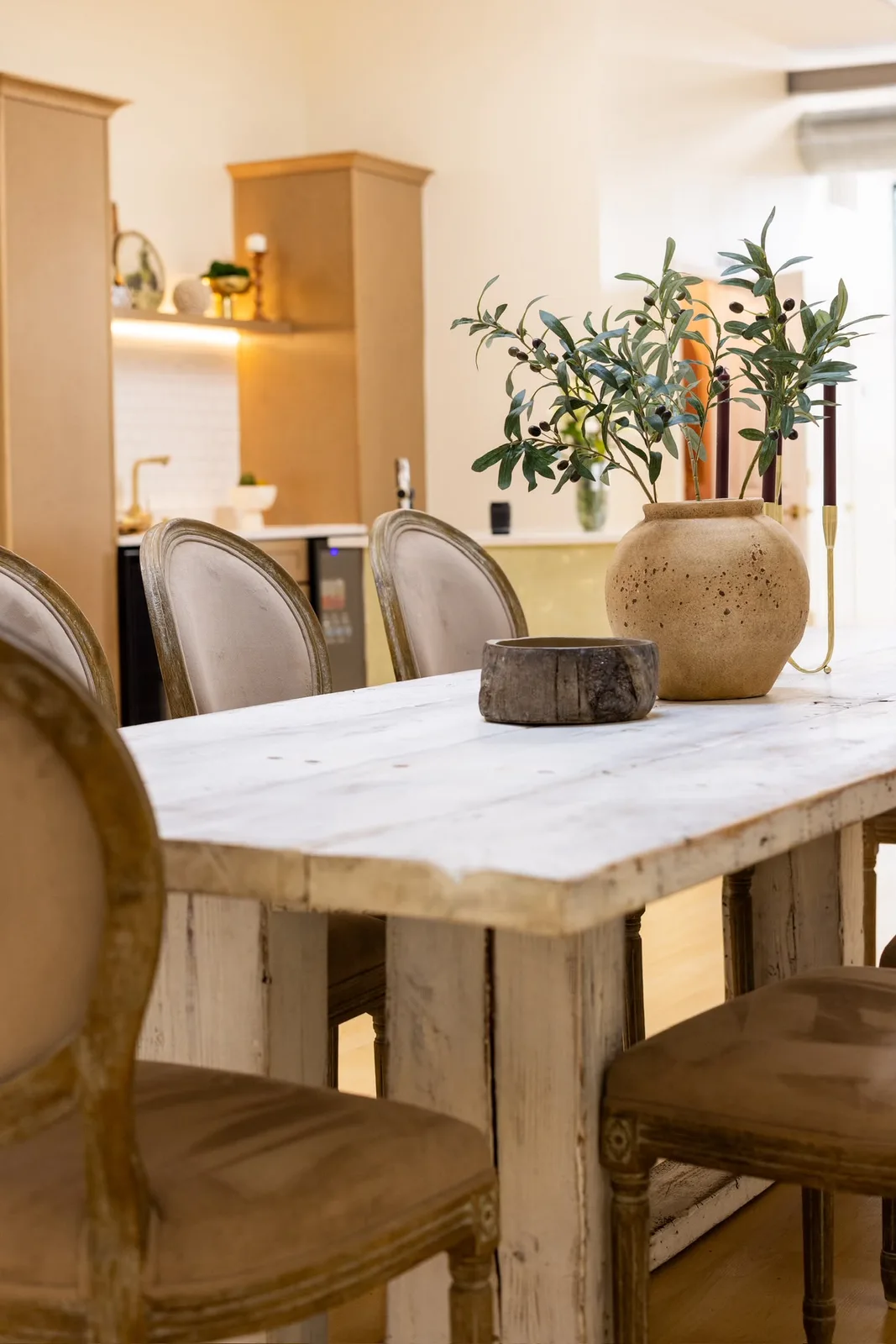 Rustic dining table with upholstered chairs and olive branches in a clay vase